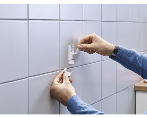 Une personne fixe un crochet adhésif sur un mur carrelé.