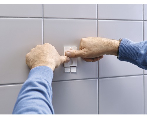 Une personne fixe un crochet adhésif sur un mur carrelé.