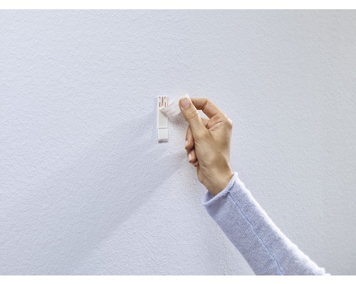 Eine Hand befestigt einen selbstklebenden Bilderhaken an einer Wand.