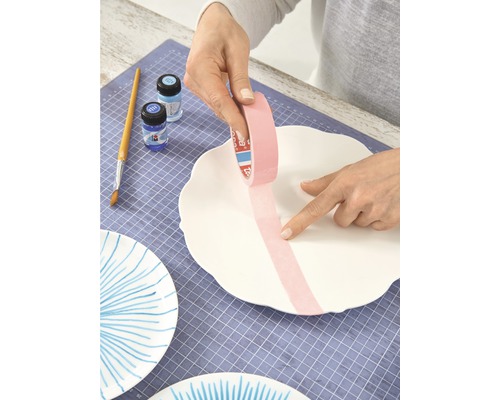 Eine Person klebt Malerkrepp auf eine weiße Keramikplatte, um sie zu dekorieren. Pinsel, Farben und weitere bemalte Platten sind sichtbar.