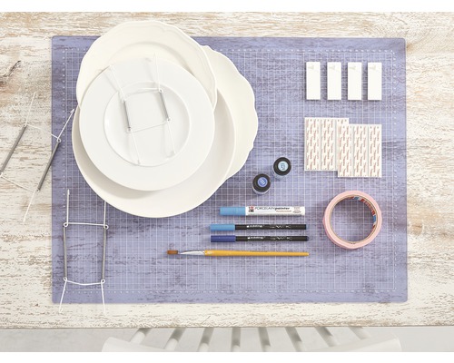 Szene mit Tellern, Tellerträgern, Porzellanfarben, Pinseln, Klebeband und Befestigungsmaterial auf einer Schneidematte