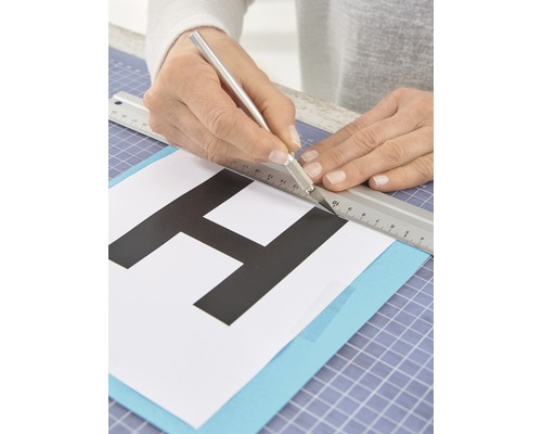 Eine Person schneidet mit einem Bastelmesser und einem Lineal einen Buchstaben aus Papier.