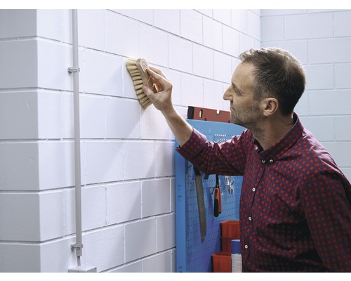 Un homme nettoie un mur de briques blanc avec une brosse. Des outils sont accrochés à un panneau mural pour outils.