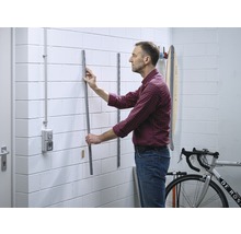 Un homme fixe un rail métallique au mur pour ranger des objets dans un garage ou une cave.