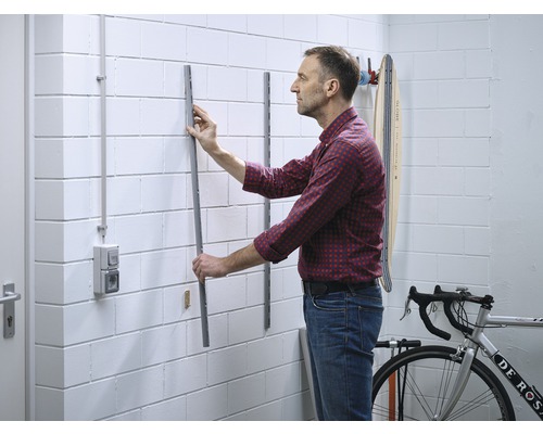 Un homme fixe un rail métallique au mur pour ranger des objets dans un garage ou une cave.
