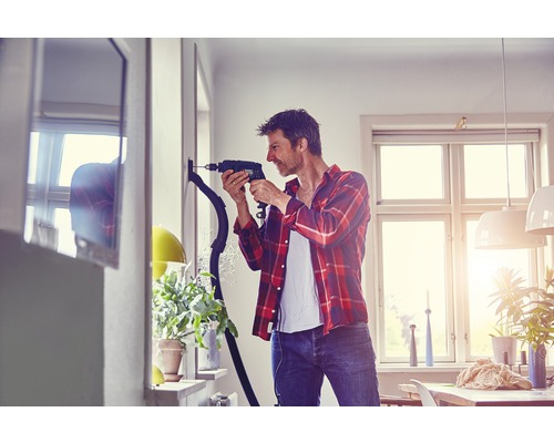 Mann bohrt mit einem Bohrgerät ein Loch in die Wand, während ein Staubsauger den Staub absaugt.