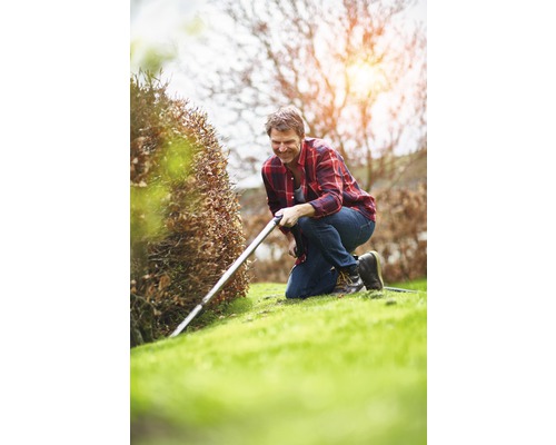 Mann kniet im Garten und schneidet eine Hecke mit einer Heckenschere.