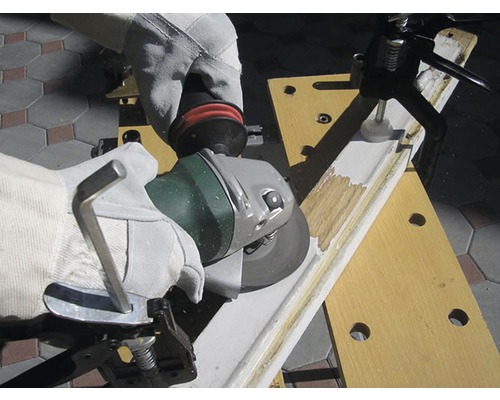 Eine Person mit Handschuhen schleift mit einem Winkelschleifer ein Werkstück ein, das mit einer Werkbank befestigt ist.