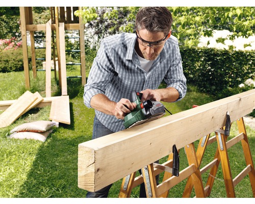Un homme rabote une planche de bois dans le jardin avec un rabot électrique.