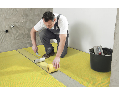 Un artisan applique des panneaux de désolidarisation sur un sol à l''aide d''un rouleau à peinture. Un seau contenant des outils se trouve à côté.