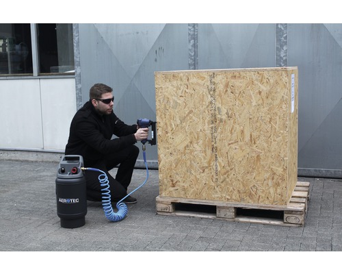 Un homme utilise un cloueur pneumatique sur une caisse en bois posée sur une palette. Un compresseur Aerotec est également visible.
