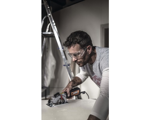 Un homme portant des lunettes de protection coupe une pièce avec une scie circulaire. Un escabeau est visible à l''arrière-plan.