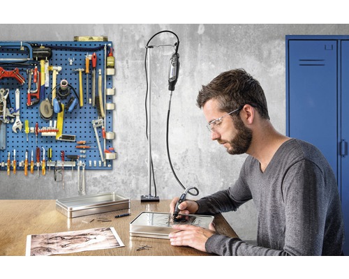 Un homme grave dans une boîte métallique avec un outil Dremel sur un bureau, mur d''outils en arrière-plan