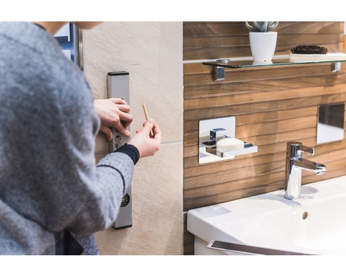 Une personne utilise un niveau à bulle et un crayon dans la salle de bain pour tracer une ligne droite sur le mur. Un lavabo, un robinet et un distributeur de savon sont visibles en arrière-plan.