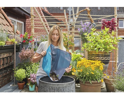 Une femme vide un sac de plantation dans un seau métallique sur un balcon avec de nombreuses plantes.