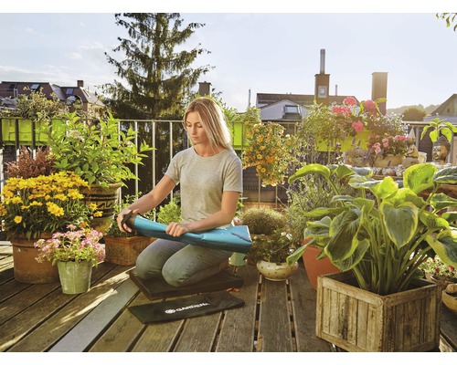 Une femme s''agenouille sur un balcon en bois et enroule un tapis de jardinage, entourée de plantes en pot et de fleurs.