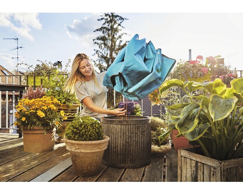 Femme vide un sac de compost dans un pot de fleurs sur un balcon, entourée de divers pots de fleurs plantés