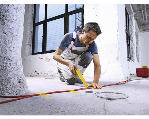 Un homme fixe un câble rouge sur un sol en béton avec du ruban adhésif.