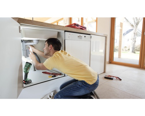 Un homme répare un drain sous un évier avec des outils et une lampe de travail dans une cuisine lumineuse.