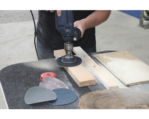 Eine Bohrmaschine mit Schleifscheibe wird verwendet, um ein Holzstück zu bearbeiten. Schleifpapier liegt bereit.