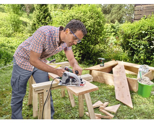 Un homme scie du bois avec une scie circulaire à main dans le jardin. Des outils et des matériaux de construction sont visibles à l''arrière-plan.