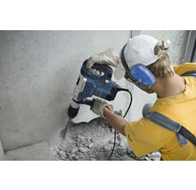 Ein Mann arbeitet mit einem Bohrhammer an einer Betonwand. Er trägt Schutzhandschuhe und einen Gehörschutz.