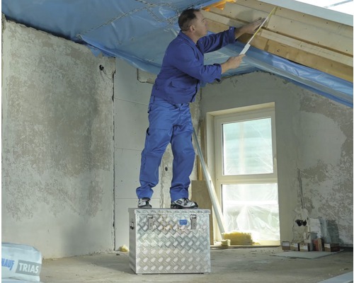 Un homme se tient sur une boîte à outils et mesure un grenier avec un mètre pliant.
