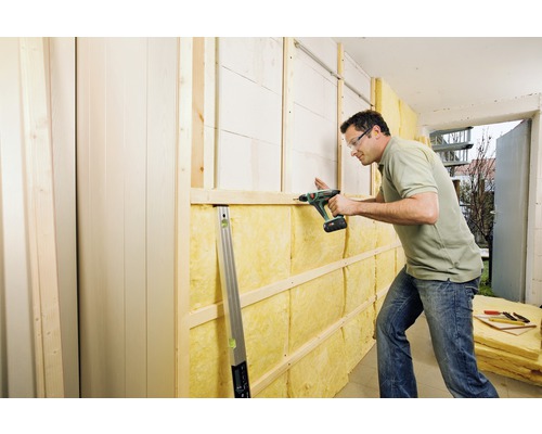 Un homme fixe de la laine isolante à un mur avec une visseuse sans fil. Cadre en bois et panneaux sont visibles.