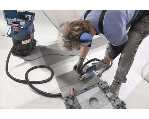 Un artisan coupe un carreau avec une meuleuse d''angle, un aspirateur industriel aspirant la poussière.