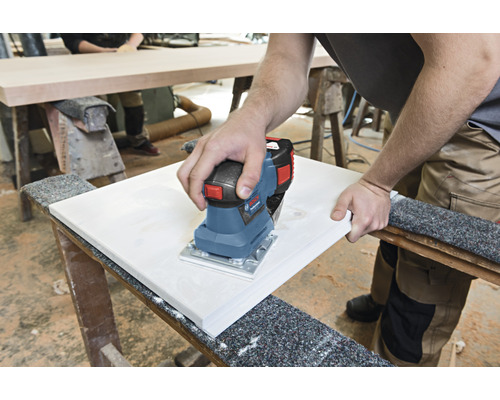 Eine Person schleift mit einem elektrischen Schwingschleifer ein Holzbrett auf einer Werkbank.