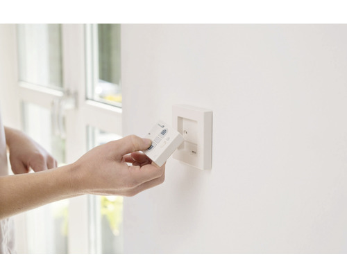 Hand hält eine Fernbedienung vor einer Wandhalterung für Leuchten.