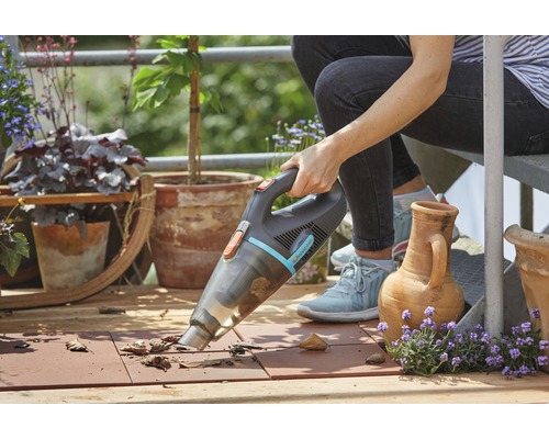 Eine Person verwendet einen Akku-Handsauger auf einem Balkon, um Blätter zu entfernen. Blumentöpfe und Pflanzen sind im Hintergrund zu sehen.