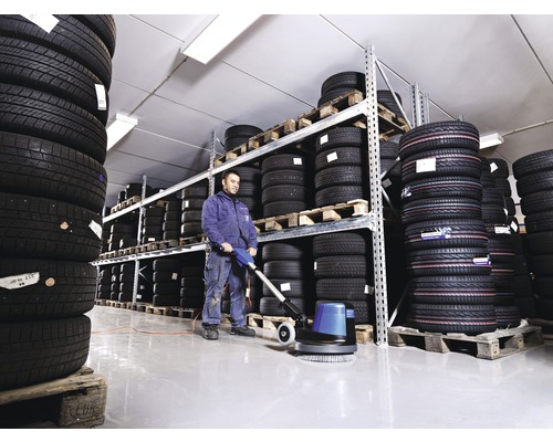 Mann reinigt Lagerhalle mit Reinigungsmaschine vor Reifenstapeln auf Paletten