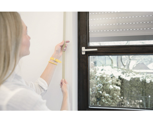 Frau misst mit einem Rollmeter die Wand neben einem Fenster mit Rollladen.