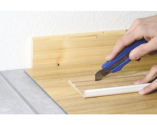 Ein Teppichmesser schneidet eine weiße Fußleiste auf einer Holzoberfläche.