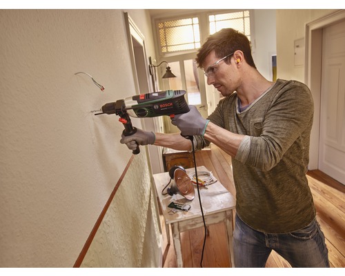 Ein Mann bohrt mit einer Schlagbohrmaschine ein Loch in eine Wand. Der Mann trägt eine Schutzbrille und Handschuhe.