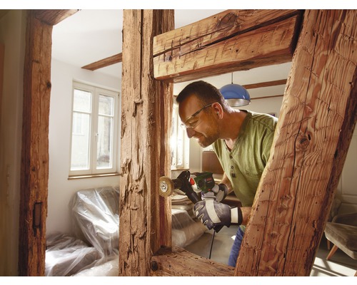 Ein Mann arbeitet mit einer Schleifmaschine an einem Holzbalken im Innenbereich.