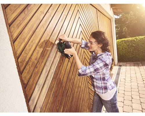 Une femme ponce une porte en bois avec une ponceuse. Projet de bricolage à l''extérieur.