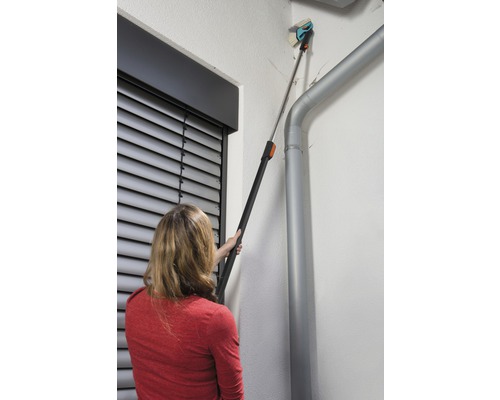 Une femme utilise une brosse télescopique pour nettoyer une façade à côté d''une gouttière.