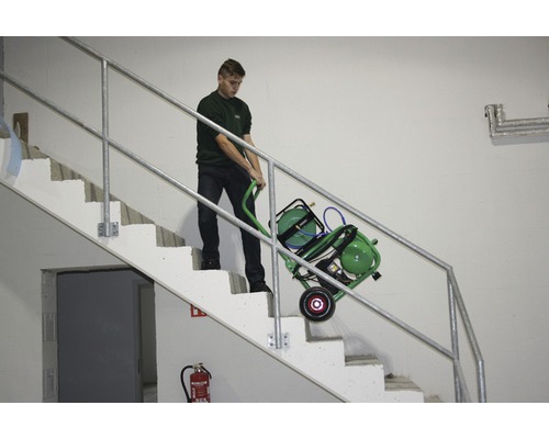 Un homme transporte un compresseur vert avec un chariot à tuyau dans un escalier.