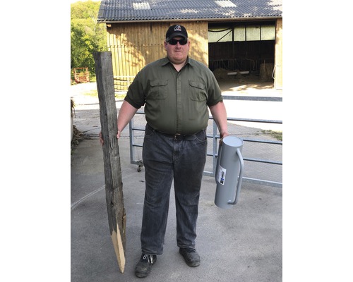 Un homme tient un poteau en bois et un vibreur de clôture devant un bâtiment d''écurie