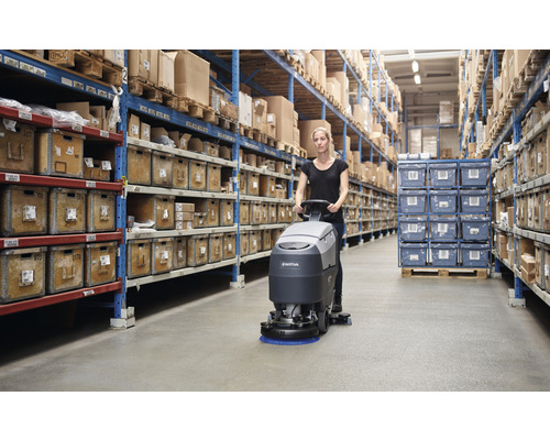 Une femme utilise une autolaveuse dans un entrepôt avec des étagères remplies de caisses.