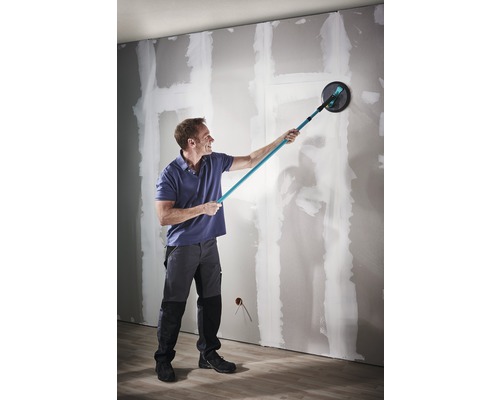 Un homme ponce un mur avec une ponceuse à plafond dans une pièce.