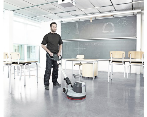 Ein Mann reinigt einen Raum mit einer Reinigungsmaschine, Tische und Stühle sind im Hintergrund zu sehen.