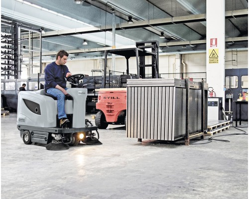 Un homme utilise une balayeuse autoportée dans un entrepôt. Un chariot élévateur et d''autres machines sont visibles en arrière-plan.