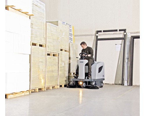 Un homme conduit une balayeuse dans un entrepôt entre des palettes de marchandises.