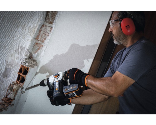 Ein Mann mit Schutzbrille und Gehörschutz arbeitet mit einem Bohrhammer an einer Ziegelwand.