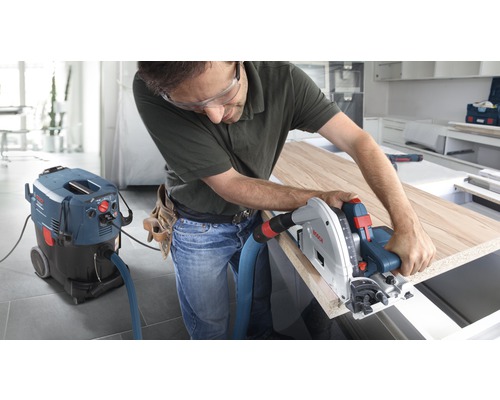 Un artisan travaille avec une scie plongeante sur une plaque de bois, raccordée à un aspirateur industriel de Bosch.