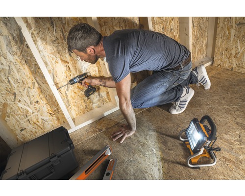 Un artisan travaille avec une visseuse sans fil dans une construction brute en bois, éclairée par un projecteur de chantier.