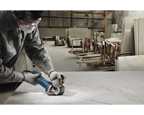 Un ouvrier portant des lunettes et un masque de protection utilise une meuleuse d''angle pour travailler une plaque de pierre dans un atelier.